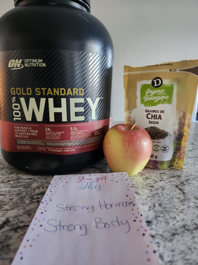 Protein powder and an apple on the counter with Strong Hormones strong body message on paper