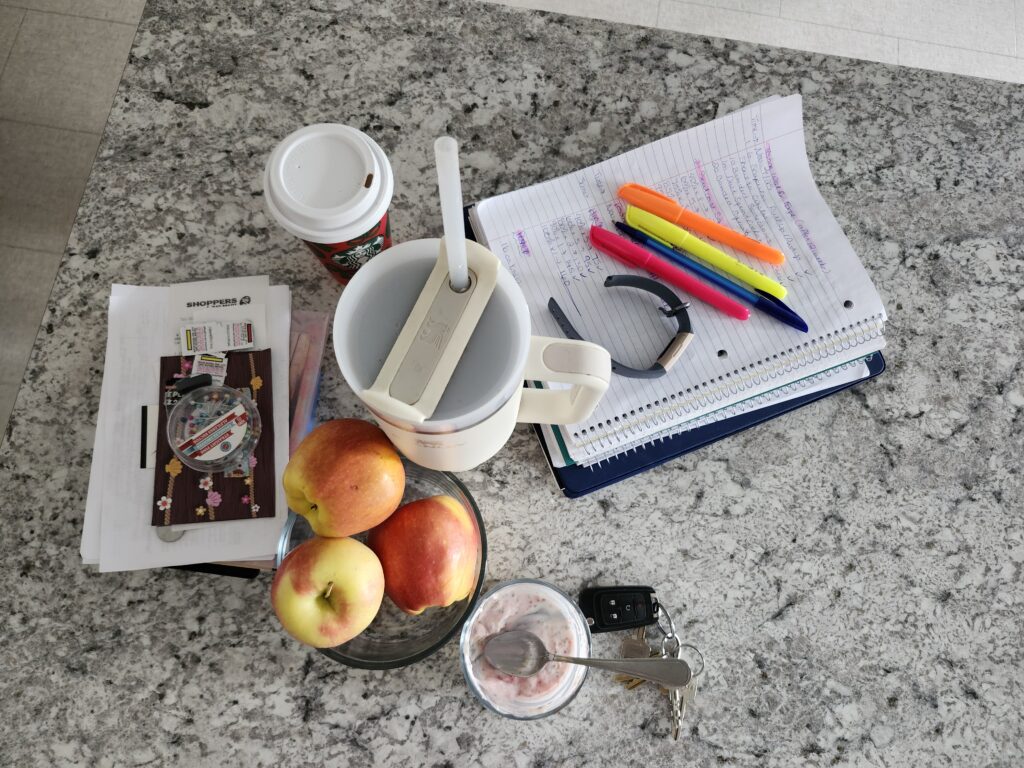 A messy counter with notebooks, highlighters, waterbottle, coffee, keys indicating a frazzled mess