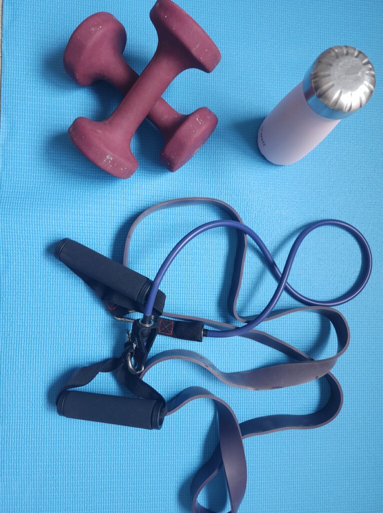 Red dumbbells, various workout bands on a blue yoga mat preparing for a home workout with a pink water bottle